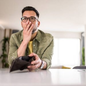 Young man sitting at home with an empty wallet. He is left without money and thinking what to do next.