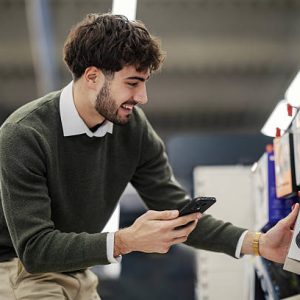Smiling young man scans the barcode on a headphones box with his smartphone while price-checking and comparing options in a modern electronics store before buying new audio gear